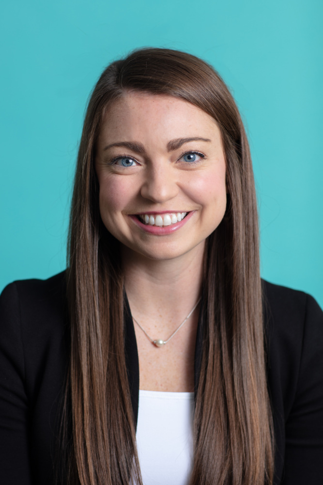 Headshot of Marie Davidson wearing a white blouse and black suit jacket on a teal background.